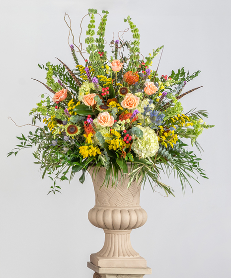 A mixture of fall flowers featuring roses, hydrangeas, protea, sunflowers, and bells of Ireland, displayed in a tan urn.