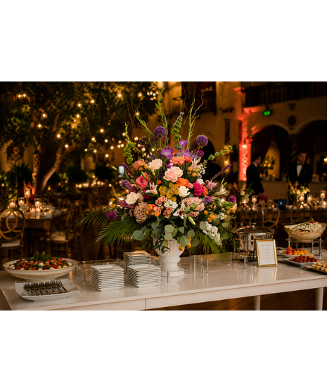 Large elevated floral arrangement featuring roses, lilies, allium, snapdragons, and mixed seasonal blooms in pink, peach, purple, and yellow tones in a white urn vase.
