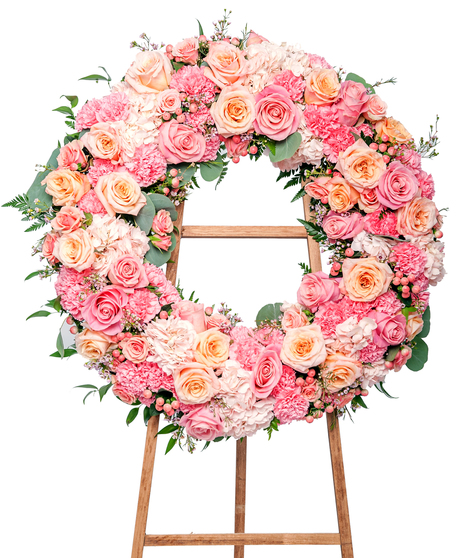 Circular floral wreath made of pastel pink and orange roses, hydrangeas, and carnations, with green leaves woven through, displayed on a wood stand against a white background.