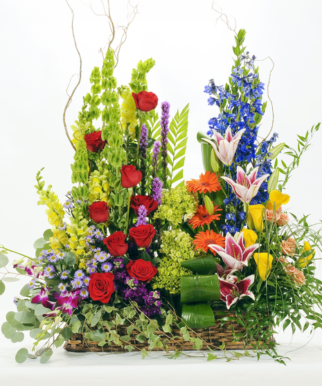A radiant floor arrangement featuring red roses, pink lilies, yellow calla lilies, orange gerbera daisies, and blue delphinium with bells of Ireland and curly willow accents.