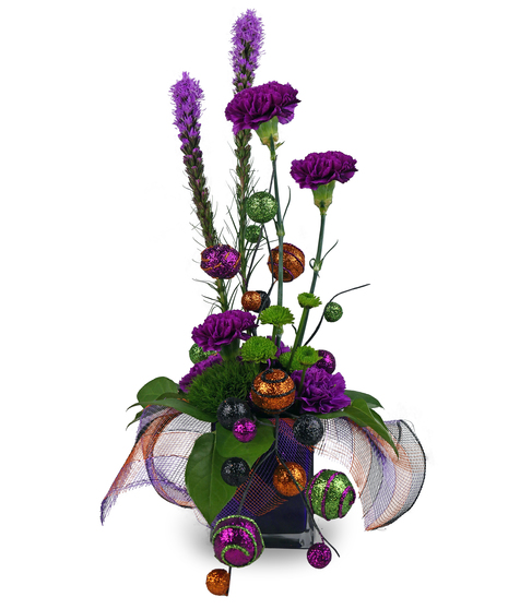 A Halloween-themed floral arrangement in a dark purple glass cube, featuring purple carnations, purple liatris, and green trick dianthus