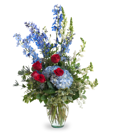A vase of roses, hydrangeas, and delphinium, mixed with greenery. Displayed in a clear vase, against a white background.