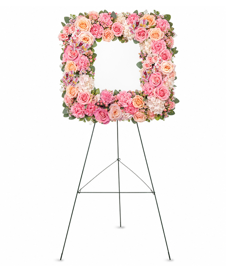 Circular floral wreath made of pastel pink and orange roses, hydrangeas, and carnations, with green leaves woven through, displayed on a wood stand against a white background.