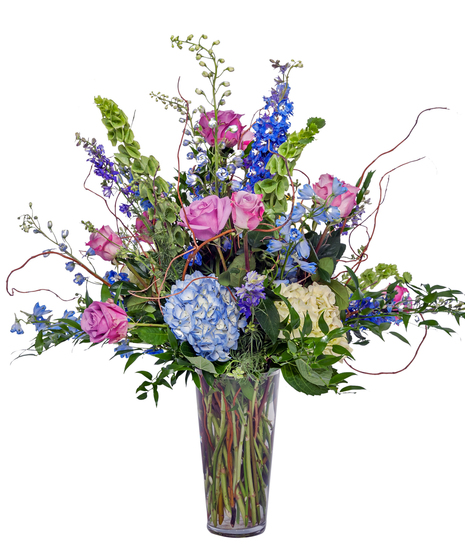 A large arrangement of pastel roses, hydrangeas, bells of Ireland, and delphinium, accented by green leaves, displayed in a clear glass vase against a white background.