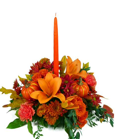 An orange taper candle in the center of a Thanksgiving floral arrangement with orange lilies, carnations, chrysanthemums, and mini pumpkins, creating a warm holiday display.
