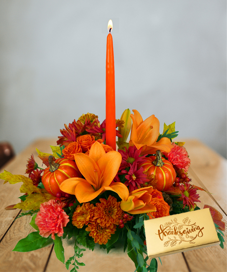 An orange taper candle in the center of a Thanksgiving floral arrangement with orange lilies, carnations, chrysanthemums, and mini pumpkins, creating a warm holiday display.