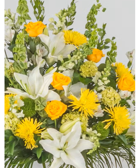 A large, lush floral arrangement with roses, white lilies, and hydrangeas, accented by green leaves, displayed in a white urn.